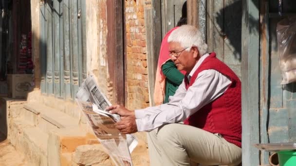 BHAKTAPUR, KATHMANDU, NEPAL - 18 de octubre de 2018 Anciano leyendo periódico en la calle. Hombre mayor con anteojos sentado en el porche de la casa. vida cotidiana, ciudad antigua oriental después del terremoto. — Vídeos de Stock