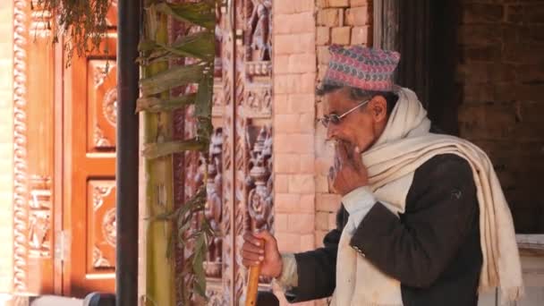 BHAKTAPUR, NEPAL - 13 PAŹDZIERNIK 2018 Starszy człowiek palący na ulicy starożytnego miasta. Etniczny mężczyzna w szaliku i okularach palący papierosa stojąc na ulicy z kamiennymi budynkami na tle. — Wideo stockowe