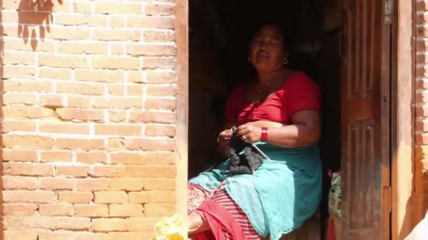BHAKTAPUR, KATHMANDU, NEPAL - 18 oktober 2018 Etnisk kvinnostickning sittande i dörröppningen. Tillfällig etnisk kvinna som sitter i öppnad dörr av rött tegelhus och stickar i solljus. — Stockvideo