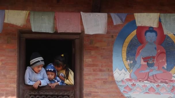 BHAKTAPUR, KATHMANDU, NEPAL - 18 de octubre de 2018 Niños en la ventana del templo envejecido. Pequeños niños étnicos en la ventana ornamental del templo budista con imagen en la pared de ladrillo con oraciones colgando de una cuerda. — Vídeos de Stock