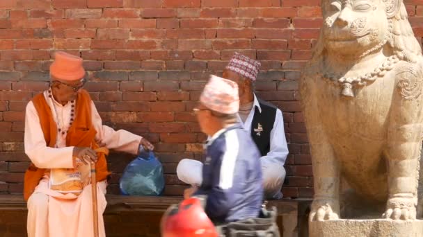 LALITPUR, NEPAL - 2018年10月7日老年人坐在石雕旁边的长椅上.在阳光下,巴丹市大街上的长椅上坐着成熟的少数民族人士.日常生活，东方古城 — 图库视频影像