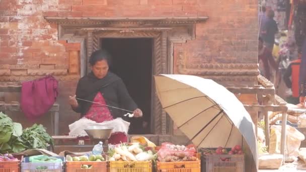 BHAKTAPUR, NEPAL - 13 OKTOBER 2018 Kvinnligt snurrande garn sittande i ett bås på gatan. Etnisk kvinna spinning tråd på gatan sitter vid stall med grönsaker till salu i solljus. — Stockvideo