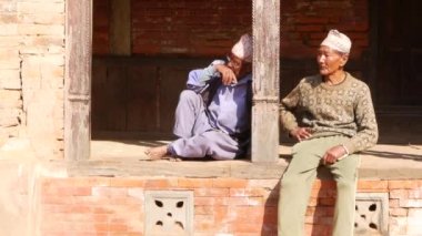 BHAKTAPUR, Katmandu, Nepal - sokakta oturan 18 Ekim 2018 yaşlı etnik adam. Çıplak ayakla ahşap terasta oturan iki yaşında etnik adam. günlük yaşam, deprem sonrası oryantal antik kenti