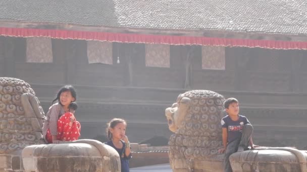 BHAKTAPUR, KATHMANDU, NEPAL - 18 de octubre de 2018 Pequeñas niñas alegres sentadas en la escultura de piedra, jugando en el patio del templo envejecido a la luz del sol. Vida cotidiana, ciudad antigua oriental después del terremoto — Vídeo de stock