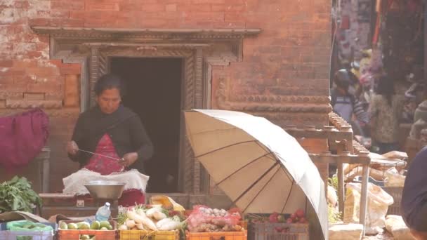 BHAKTAPUR, NEPAL - 13 OKTOBER 2018 Vrouw spinnen garen zitten op stal op straat. Etnische vrouw spinnen draad op straat zitten op stal met groenten te koop in zonlicht. — Stockvideo