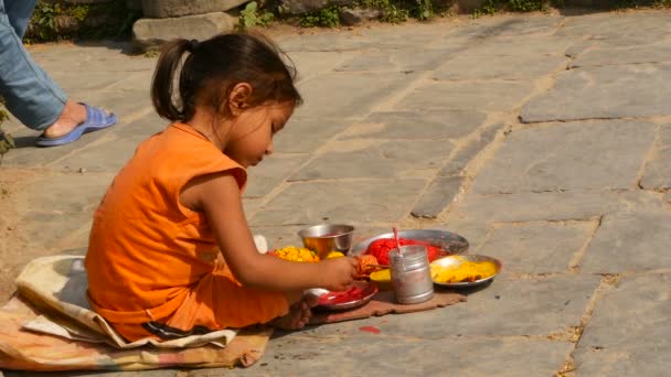 KATHMANDU, NEPAL - 8. OKTOBER 2018 Seitenansicht eines kleinen ethnischen Mädchens, das verschiedene religiöse Attribute verkauft und auf dem Steinpflaster des Tempels sitzt. ghats in Paschupatinath. Hindu traditioneller Brauch. — Stockvideo