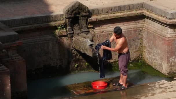 BHAKTAPUR, NEPAL - 13 October 2018寺院の壁の近くで洗濯する男。通りのレンガの壁に高齢者の石のクレーンからの水で髪や服を洗う若い男のビュー。カトマンズの街並み. — ストック動画