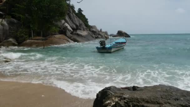 Båt på land nær bølgende hav. Tradisjonell båt på sandkysten nær praktfullt bølgende hav på solskinnsdag på tropisk eksotisk Koh Phangan i Thailand. Fullmåne party strand Haad Rin. Paradis – stockvideo