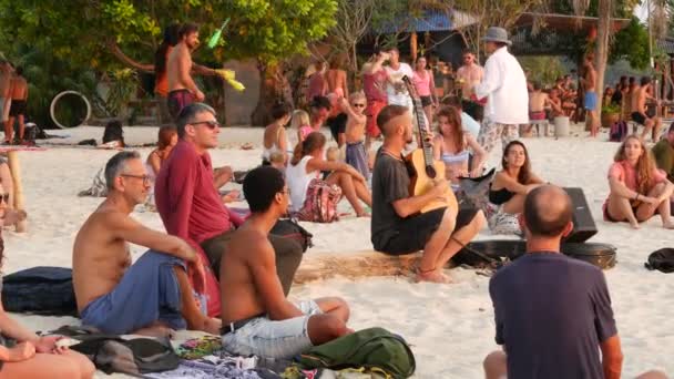 PHANGAN, ThAILAND - 23 e.Kr. Zen Beach 2019. Leende ung kille spelar gitarr på en tropisk paradisets sommarkust vid solnedgången omgiven av många människor. Musiksylt, frihetskoncept, social fritid — Stockvideo