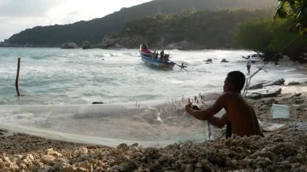 KOH PHANGAN, TAILÂNDIA - 11 JANEIRO 2019 Pescador étnico isca pendurada na rede. Vista lateral do homem sem camisa étnica pendurado pequeno peixe na rede enquanto sentado na costa perto do mar — Vídeo de Stock