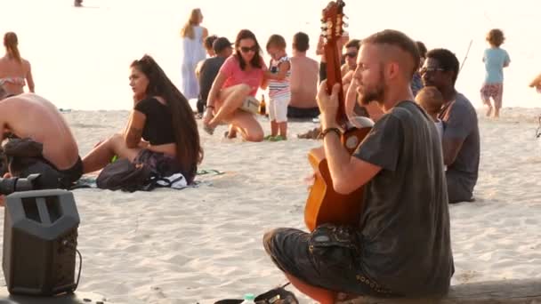 PHANGAN, TAILÂNDIA - 23 DE FEVEREIRO DE 2019 Zen Beach. Jovem sorridente toca guitarra em uma costa de verão paradisíaca tropical ao pôr-do-sol cercado por muitas pessoas. Engarrafamento musical, conceito de liberdade, lazer social — Vídeo de Stock