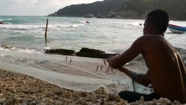 KOH PHANGAN, THAILAND - 11. červen 2019 Etnický rybář visící návnadu na síti. Boční pohled na etnický muž bez košile zavěšení malé ryby na síti, zatímco sedí na břehu u moře — Stock video