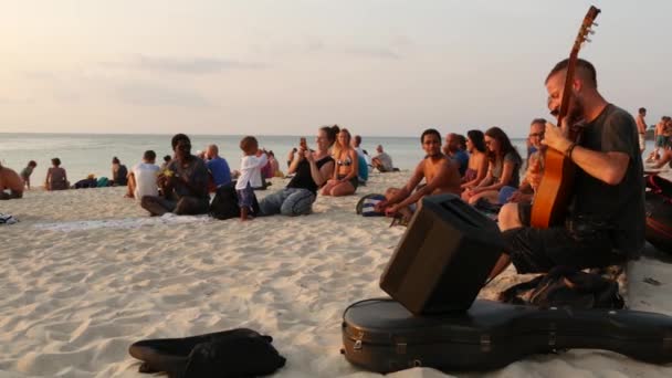 PHANGAN, TAILÂNDIA - 23 DE FEVEREIRO DE 2019 Zen Beach. Jovem sorridente toca guitarra em uma costa de verão paradisíaca tropical ao pôr-do-sol cercado por muitas pessoas. Engarrafamento musical, conceito de liberdade, lazer social — Vídeo de Stock