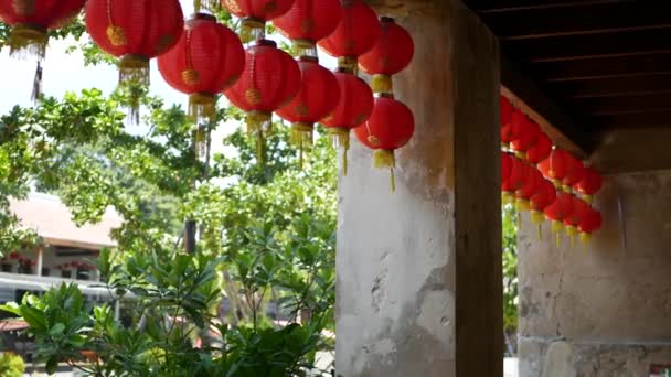 Paper lanterns on shabby building. Red paper lanterns hanging on ceiling of weathered concrete temple building on sunny day between juicy greenery in oriental country. traditional decoration — Stock Video