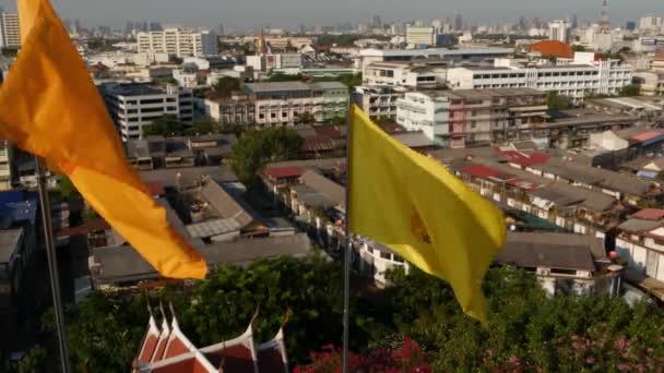 Городской пейзаж современного восточного города. Вид на крыши домов на улицах величественного Бангкока с Золотой горы во время заката солнца. Желтый королевский флаг и символ Таиланда, раскачивающегося на ветру — стоковое видео