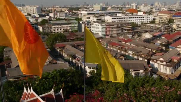 近代的なオリエンタルタウンの街並み。日没時にゴールデンマウント寺院から壮大なバンコクの通りに家の屋根のビュー。黄色の王室の旗と風に揺るぐタイのシンボル — ストック動画