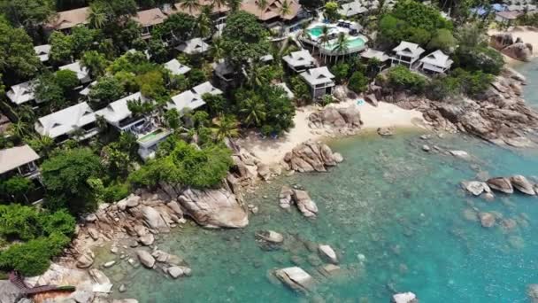 Małe domki na tropikalnej wyspie. Małe przytulne bungalowy położone na brzegu wyspy Koh Samui w pobliżu spokojnego morza w słoneczny dzień w Tajlandii. Wulkaniczne skały i klify drona widok z góry. — Wideo stockowe