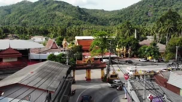 Samochody jeżdżą wzdłuż drogi w pobliżu bram świątynnych. Pojazdy jeżdżąc wzdłuż asfaltowej drodze przez małe miasteczko w pobliżu bram tradycyjnej orientalnej świątyni w dzielnicy Lamai na wyspie Koh Samui w Tajlandii. Widok Drone. — Wideo stockowe