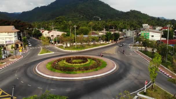 Coches montando en la rotonda en la pequeña ciudad cerca de la selva. Vehículos montados en la intersección circular de la carretera principal en medio de la pequeña ciudad contra la montaña y la selva en la isla de Koh Samui Tailandia. Vista del dron — Vídeos de Stock