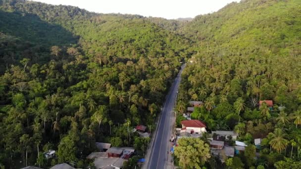 Aszfaltozott út a településen és a dzsungelen át. Aszfaltozott út megy keresztül kis tipikus település és zöld trópusi esőerdő Koh Samui szigeten. Hegyek a láthatáron. Falusi élet Drónnézet. — Stock videók