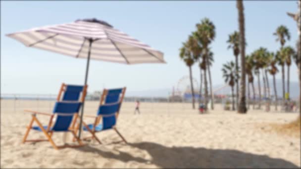 California beach defocused, two empty blue deck chairs, striped umbrella near pier in Santa Monica pacific ocean resort. Summertime relax lounge dreaming atmosphere and palm trees, Los Angeles CA USA — Stock Video