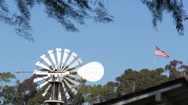 Klasický retro větrný mlýn, lopatkový rotor a americká vlajka proti modré obloze. Ročník vodní čerpadlo větrné turbíny, generátor energie na dobytčí farmě nebo zemědělské farmě. Venkovský symbol divokého západu, venkovské předměstí — Stock video
