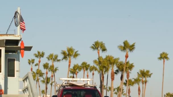 Ikonická retro dřevěná strážní věž a baywatch červené auto. Životní bóje, americká státní vlajka a palmy proti modré obloze. Summertime california estetic, Santa Monica beach, Los Angeles, CA USA — Stock video