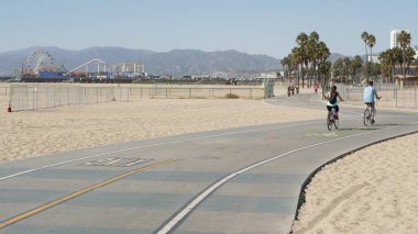 Santa Monica, LOS ANGELES CA USA - 28 OCT 2019: Kaliforniya yazları plaj estetiği, insanlar bisiklet yolunda yürüyor ve bisiklete biniyor. İskeledeki lunapark ve palmiyeler. Amerikan Pasifik Okyanusu tatil köyü.