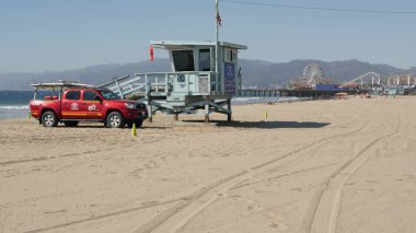 Santa Monica, LOS ANGELES CA ABD - 28 OCT 2019 Kaliforniya yaz plaj estetiği. İkonik mavi ahşap gözetleme kulesi, kumlu sahilde kırmızı cankurtaran arabası. Eğlence parkı ve rıhtımdaki eğlence.