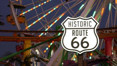 Santa Monica, LOS ANGELES, ABD - 28 OCT 2019: Iconic yol işareti parlıyor, tarihi rota 66. Meşhur California sembolü, Pasifik Okyanusu kıyısındaki rıhtım. Eğlence parkında aydınlatılmış şenlikli dönme dolap..