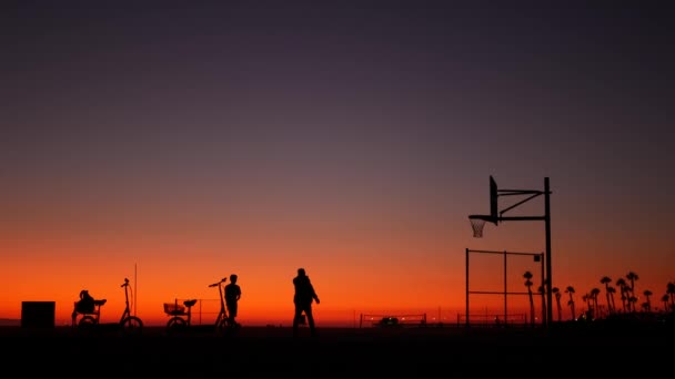 California summertime dusk beach aesthetic, pink sunset. Unrecognizable silhouettes, people play game with ball on basketball court. Newport ocean resort near Los Angeles CA USA. Purple sky gradient — Stock Video