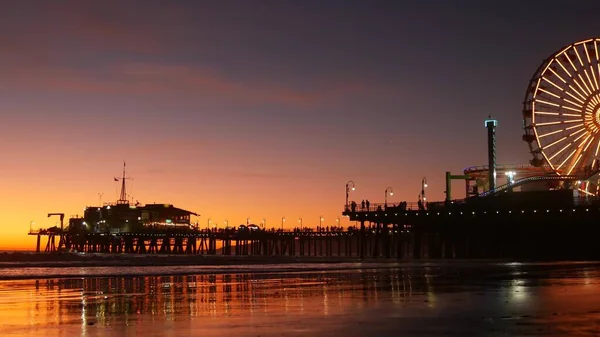 Twilight waves against classic illuminated ferris wheel, amusement park on pier in Santa Monica pacific ocean beach resort. Summertime iconic symbol of California glowing in dusk, Los Angeles, CA USA