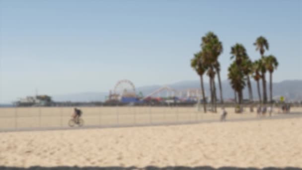 California spiaggia estetica, le persone vanno in bicicletta su una pista ciclabile. Sfondo sfocato e sfocato. Parco divertimenti su pontile e palme a Santa Monica località americana del Pacifico, Los Angeles CA USA — Video Stock