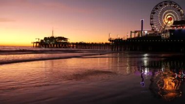 Klasik ışıklı dönme dolaba karşı alacakaranlık dalgaları, Santa Monica Pasifik Okyanusu sahilindeki rıhtımda eğlence parkı. Kaliforniya 'nın simgesel simgesi. Alacakaranlıkta parlıyor, Los Angeles, CA USA.