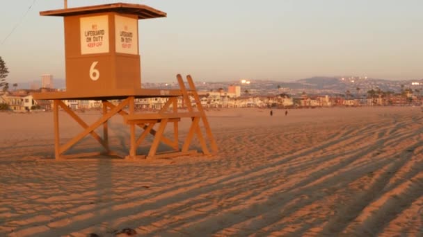 Iconic retro wooden orange lifeguard watch tower on sandy california pacific ocean beach illuminated by sunset rays. Private holiday houses and mountains on horizon. Newport resort aesthetic, USA — Stock Video