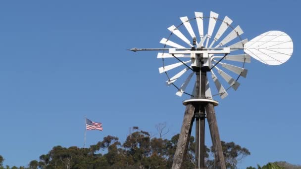 Klasyczny wiatrak retro, wirnik łopatkowy i flaga USA przeciwko błękitnemu niebu. Zabytkowa pompa wody turbina wiatrowa, generator energii na ranczu inwentarza żywego lub w gospodarstwie rolnym. Wiejski symbol dzikiego zachodu, rustykalne przedmieścia — Wideo stockowe