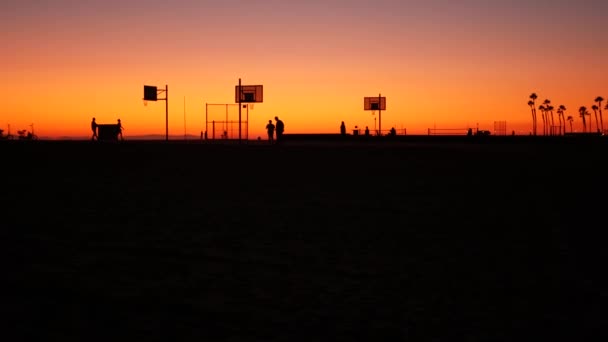 Калифорнийские летние сумерки, эстетика пляжа, розовый закат. Неузнаваемые силуэты, люди играют с мячом на баскетбольной площадке. Океанский курорт Ньюпорт недалеко от Лос-Анджелеса, США. Пурпурный градиент неба — стоковое видео