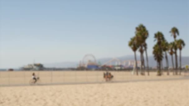 California beach aesthetic, people ride cycles on a bicycle path. Blurred, defocused background. Amusement park on pier and palms in Santa Monica american pacific ocean resort, Los Angeles CA USA — Stock Video