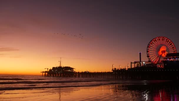 Olas del crepúsculo contra la clásica noria iluminada, parque de atracciones en el muelle de Santa Mónica complejo de playa del océano Pacífico. Símbolo icónico de verano de California brillando al atardecer, Los Ángeles, CA USA. — Vídeo de stock