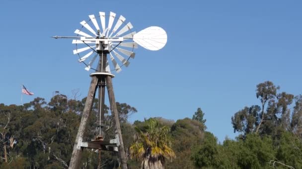 Classic retro windmill, bladed rotor and USA flag against blue sky. Vintage water pump wind turbine, power generator on livestock ranch or agricultural farm. Rural symbol of wild west, rustic suburb — Stock Video