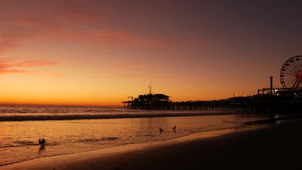 Twilight waves against classic illuminated ferris wheel, amusement park on pier in Santa Monica pacific ocean beach resort. Summertime iconic symbol of California glowing in dusk, Los Angeles, CA USA. — Stock Video