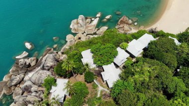 Tropikal adada küçük evler. Güneşli Tayland 'da güneşli bir günde, Koh Samui adasının kıyısındaki küçük, rahat bungalovlar. Volkanik kayalar ve uçurumlar insansız hava aracı görüntüsü