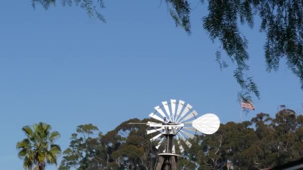 Classic retro windmill, bladed rotor and USA flag against blue sky. Vintage water pump wind turbine, power generator on livestock ranch or agricultural farm. Rural symbol of wild west, rustic suburb — Stock Video