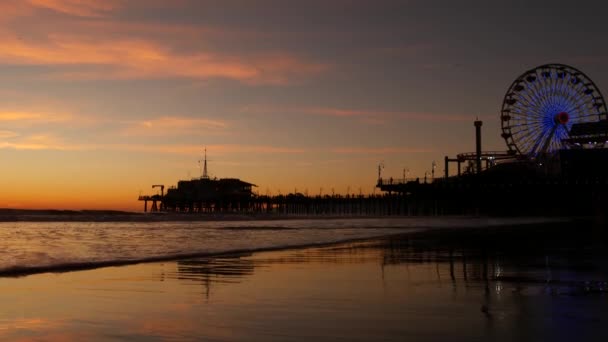 Twilight golven tegen klassieke verlichte reuzenrad, pretpark op pier in Santa Monica pacific oceaan strand resort. Summertime iconisch symbool van Californië gloeiend in de schemering, Los Angeles, CA Verenigde Staten. — Stockvideo