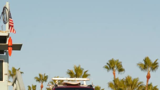 Iconic retro wooden lifeguard watch tower and baywatch red car. Life buoy, american state flag and palm trees against blue sky. Summertime california aesthetic, Santa Monica beach, Los Angeles, CA USA — Stock Video