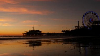 Klasik ışıklı dönme dolaba karşı alacakaranlık dalgaları, Santa Monica Pasifik Okyanusu sahilindeki rıhtımda eğlence parkı. Kaliforniya 'nın simgesel simgesi. Alacakaranlıkta parlıyor, Los Angeles, CA USA.