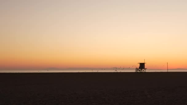 Summertime travel concept. Dark silhouette, iconic retro wooden lifeguard watch tower against sunset orange sky. Contrast watchtower outline, california pacific ocean beach twilight aesthetic, CA USA — Stock Video