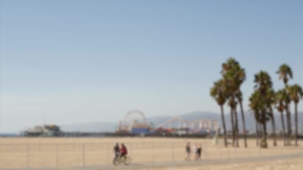 Калифорнийская пляжная эстетика, люди ездят на велосипеде по дорожке. Размытый, размытый фон. Парк аттракционов на пирсе и пальмах в Санта-Монике, США — стоковое видео