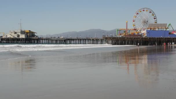 Oceaan golven en zandige Californië strand, klassieke reuzenrad in pretpark op pier in Santa Monica pacific oceaan resort. Summertime iconische weergave, symbool van Los Angeles, CA USA. Reisconcept — Stockvideo