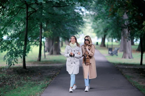 秋のファッションパークの女の子2ルック コートで2人の魅力的なファッショナブルなガールフレンドが都市公園を歩く — ストック写真
